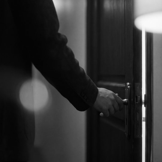 A hand reaching for a door handle in a dark hallway, warm light leaking through