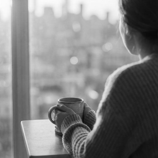 A person sitting alone at a small table by a window at sunrise, hands around a coffee mug